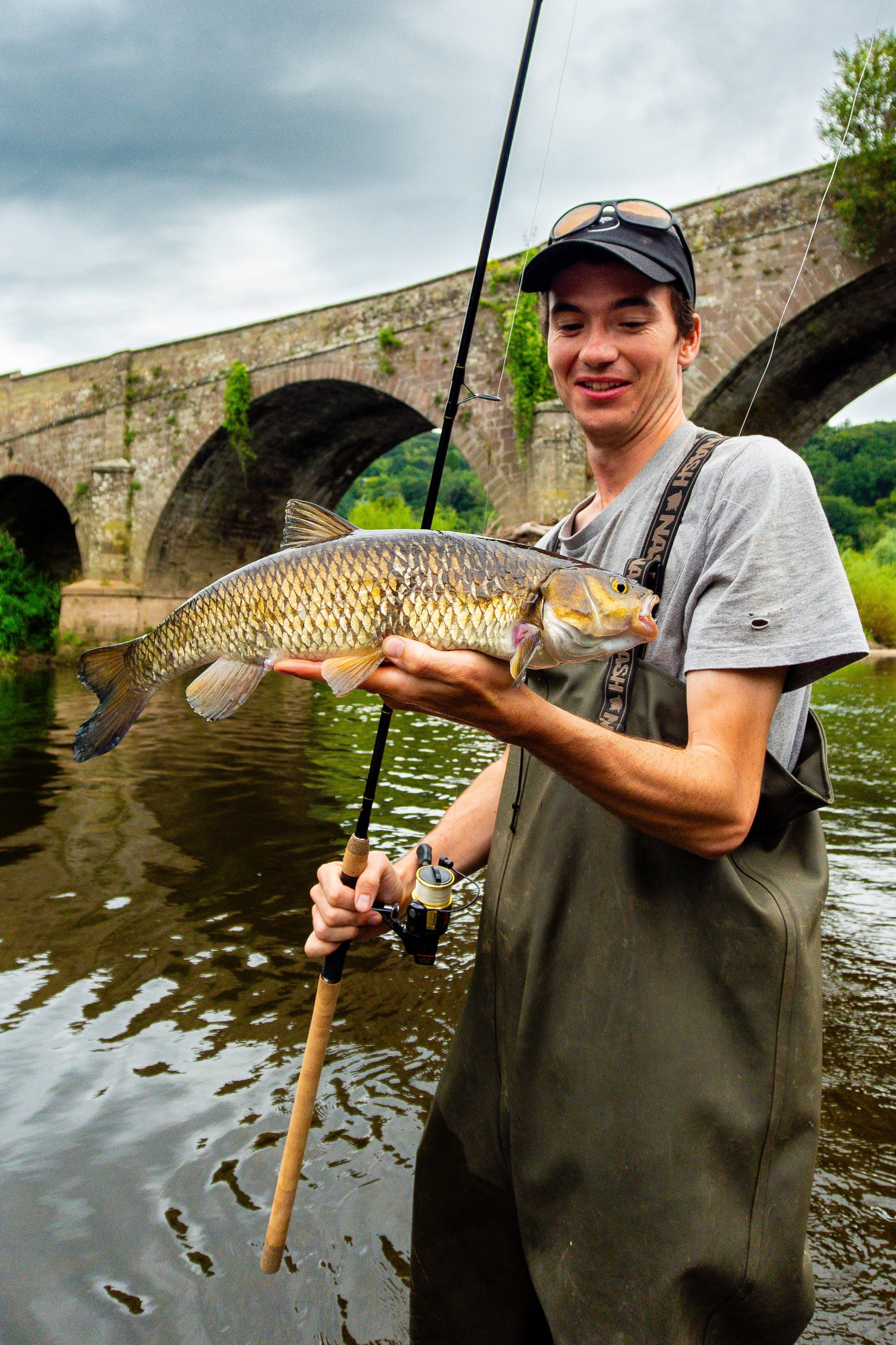 Man holding a large fish by a river with a stone bridge in the background